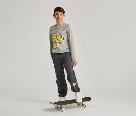 Chlapec v šedej mikine a nohaviciach so žltými smajlíkmi stojí na skateboarde pred bielym pozadím.