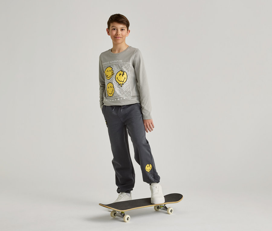 Chlapec v šedej mikine a nohaviciach so žltými smajlíkmi stojí na skateboarde pred bielym pozadím.