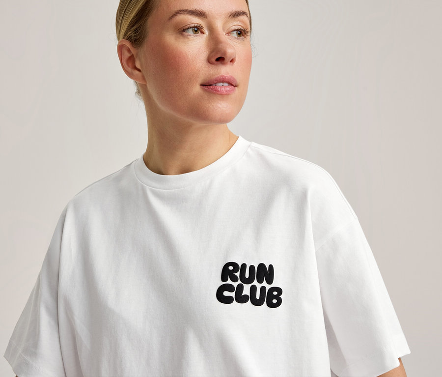 Žena má na sebe biele športové tričko s nápisom „RUN CLUB“ na hrudi.