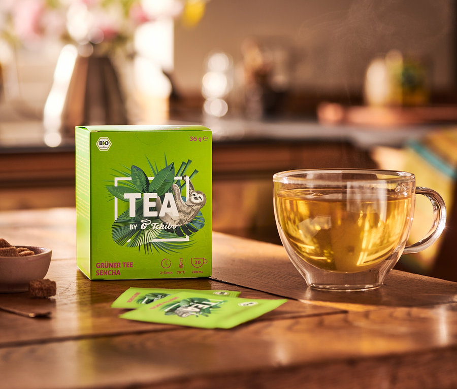 Na drevenom stole stojí balenie BIO TEA by Tchibo Sencha 8 x 20 x 1,8 g, miska s cukrom a šálka čaju.