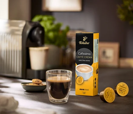 Na stole stojí kávovar, pohár s kávou a balenie Caffè Crema jemná – 120 kapsúl.
