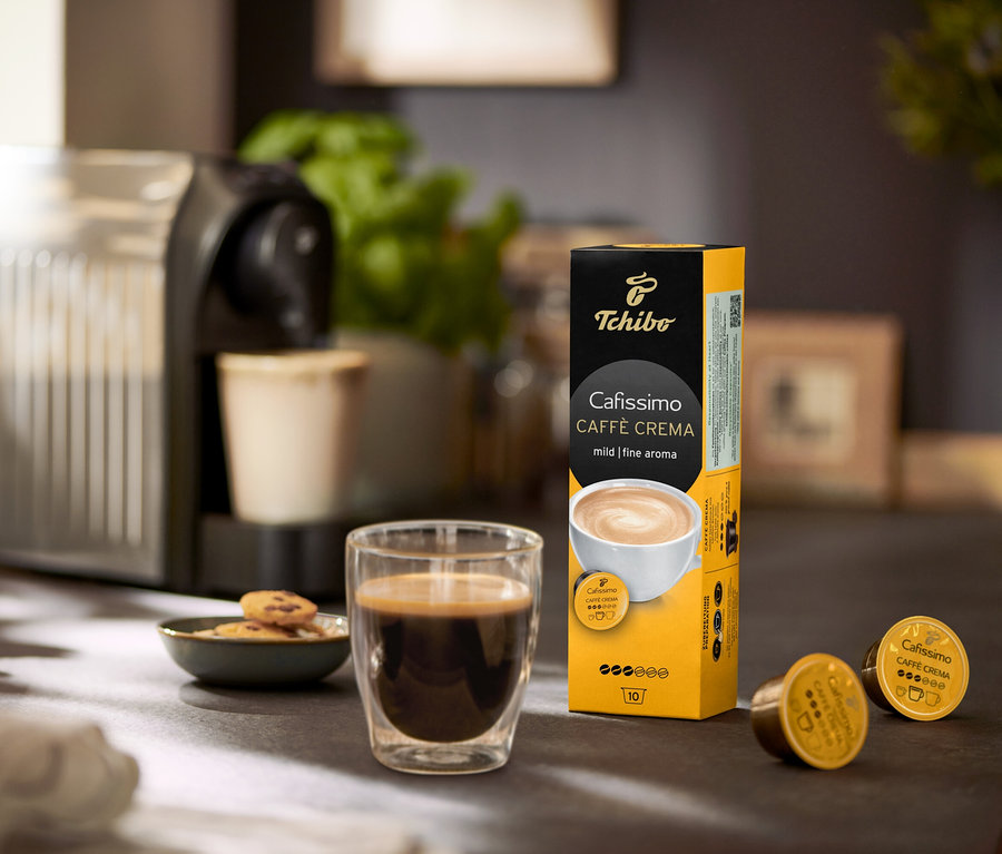 Na stole stojí kávovar, pohár s kávou a balenie Caffè Crema jemná – 120 kapsúl.