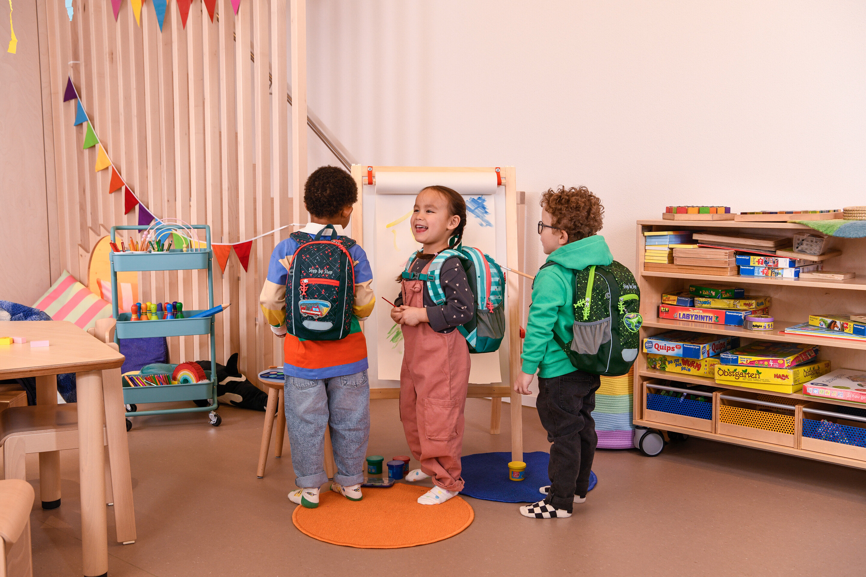 Tri deti stoja v miestnosti s batohmi a maľujú na plátno.