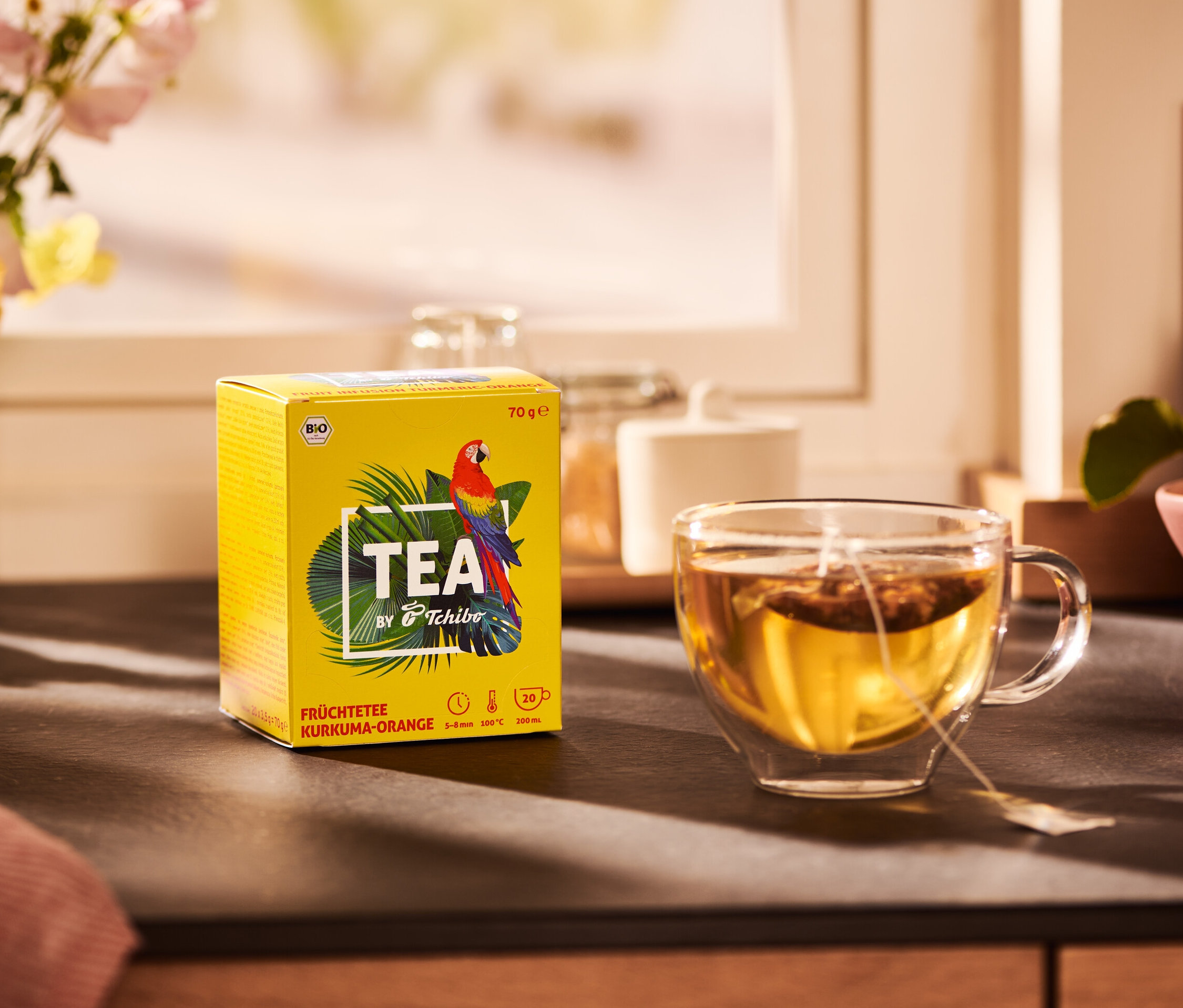 Žltá krabička čaju TEA by Tchibo BIO ovocný čaj s príchuťou kurkumy a pomaranča a šálka čaju.