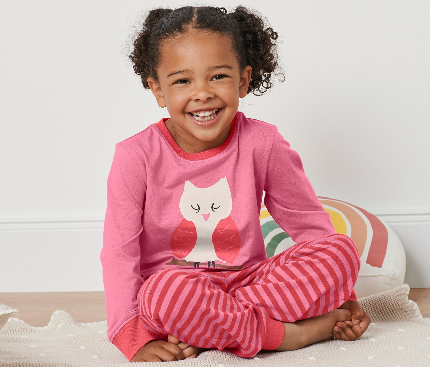 Dievča sedí na podložke v ružovo-červenom pyžame s motívom sovy.