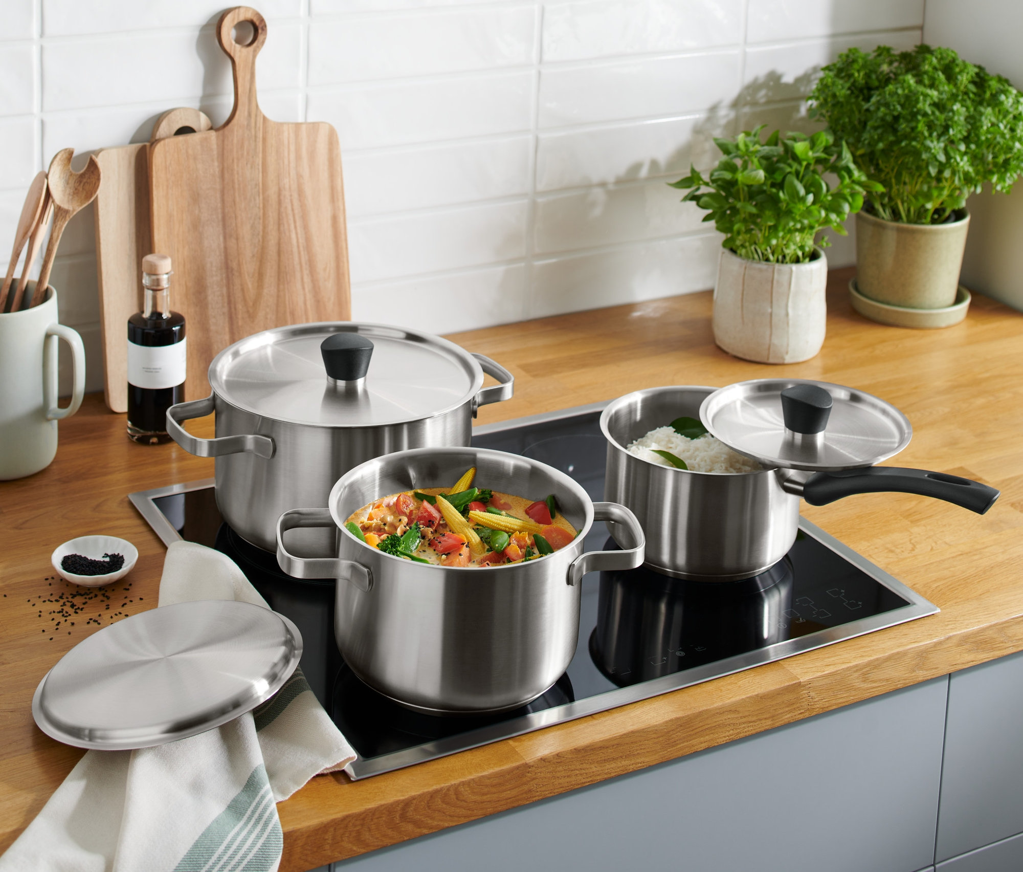 Súprava hrncov z ušľachtilej ocele stojí na indukčnej doske, jeden so zeleninou, jeden s ryžou.