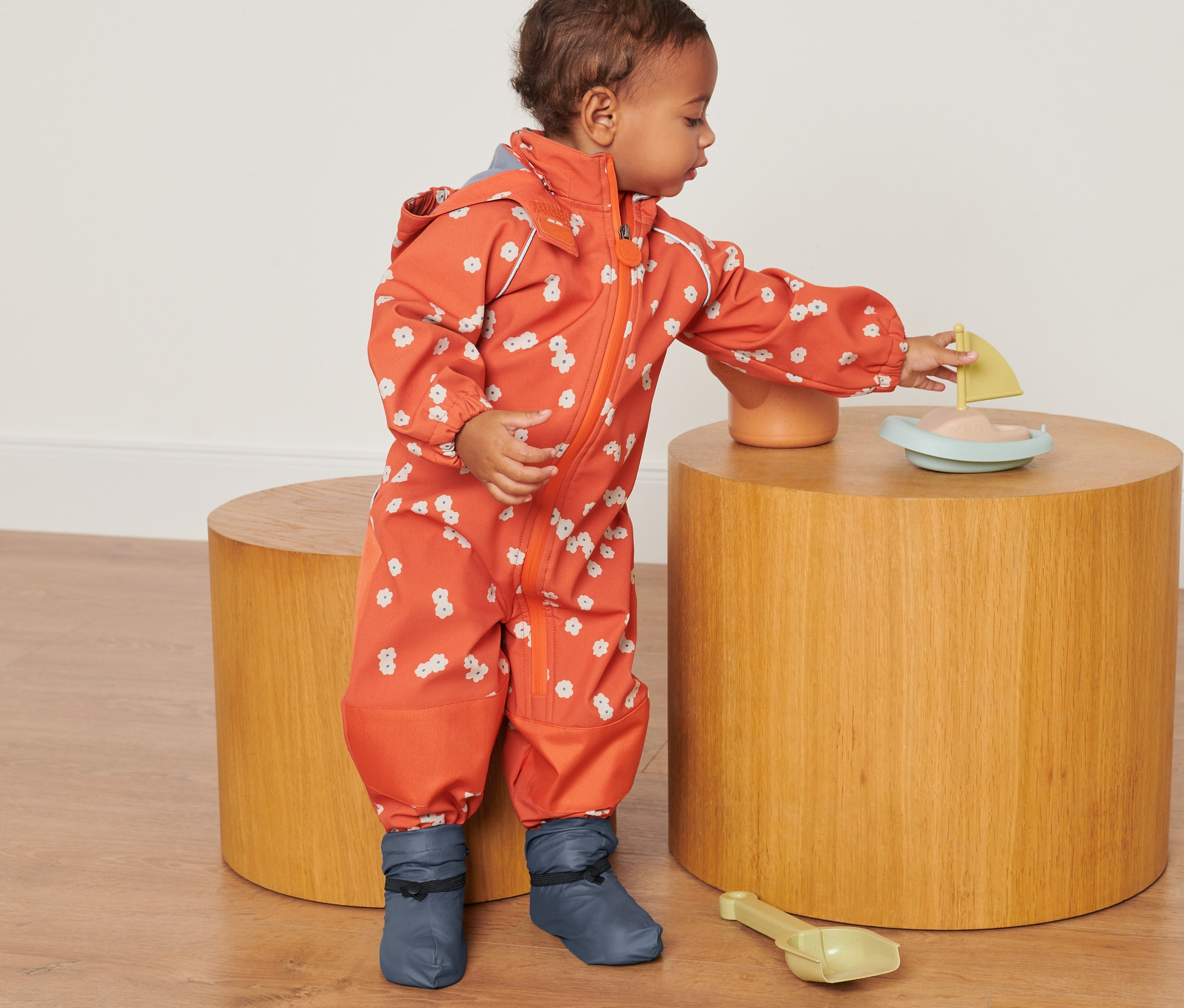 Dieťa v oranžovom overale s kvetinovým vzorom sa hrá s hračkou na drevenom stolíku a má na sebe Návleky do dažďa.