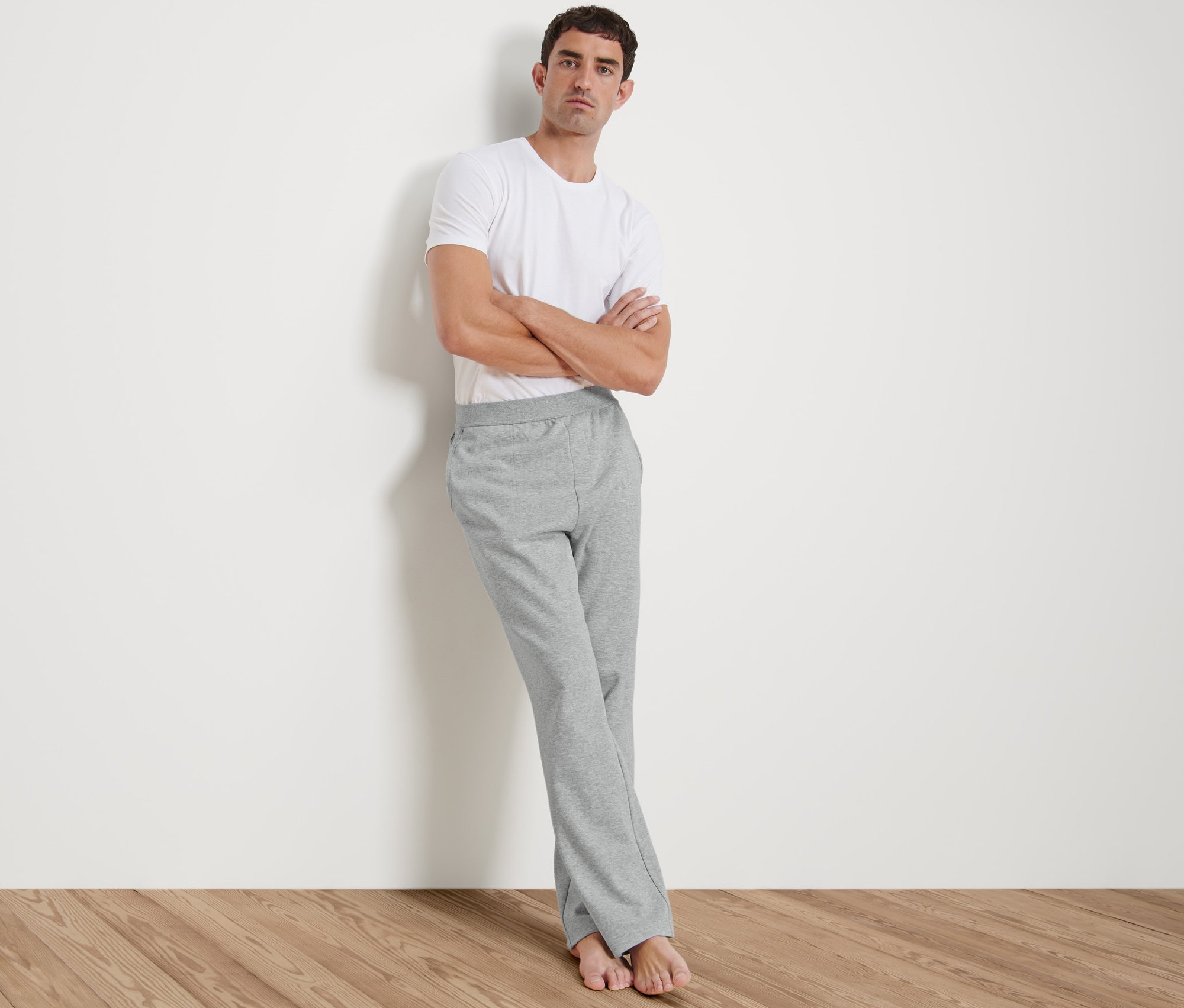 Muž opretý o stenu má na sebe biele tričko a sivé domáce nohavice.