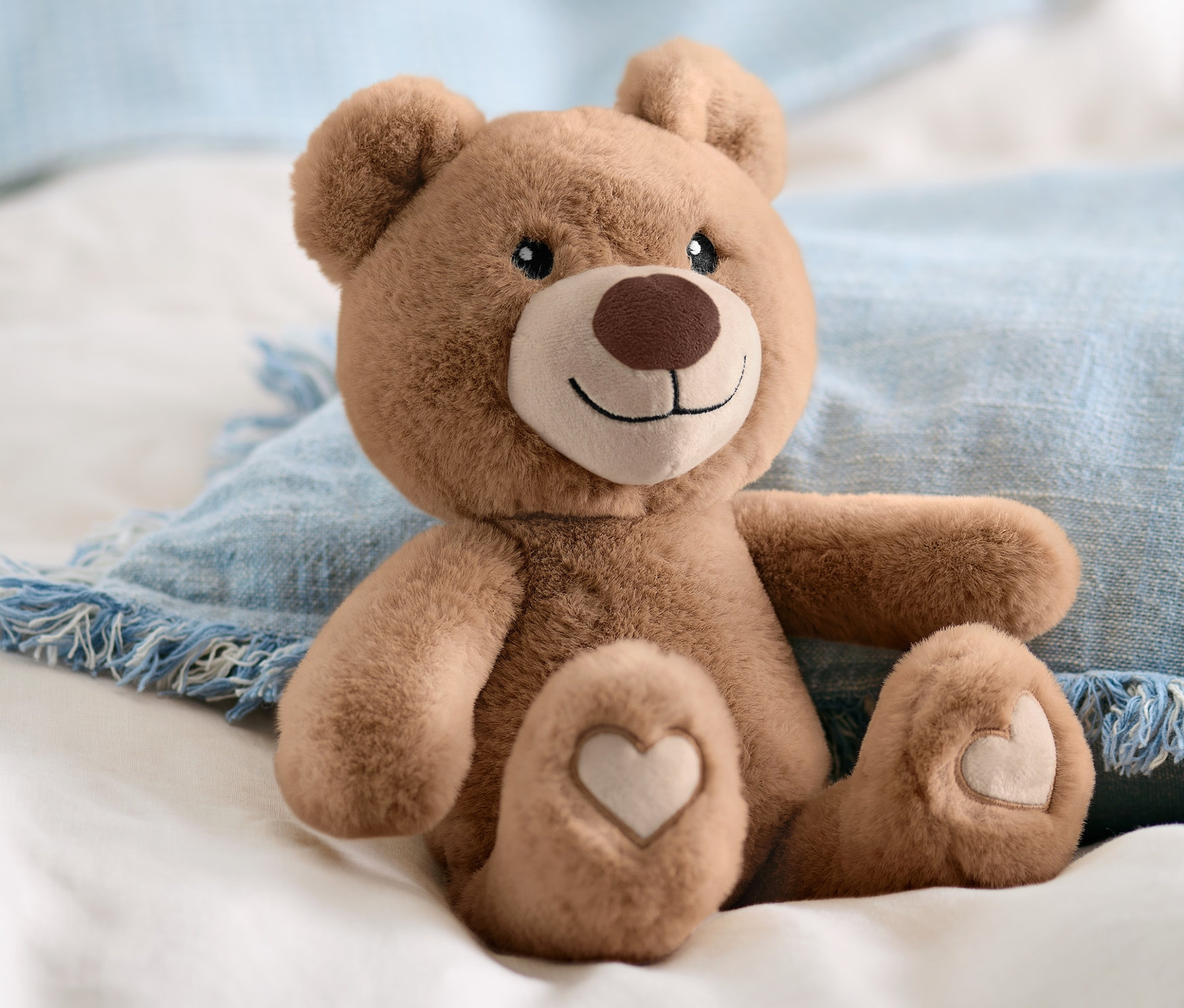 Hnedé plyšové zvieratko »Bär« sedí na bielej posteľnej bielizni a modrom vankúši.