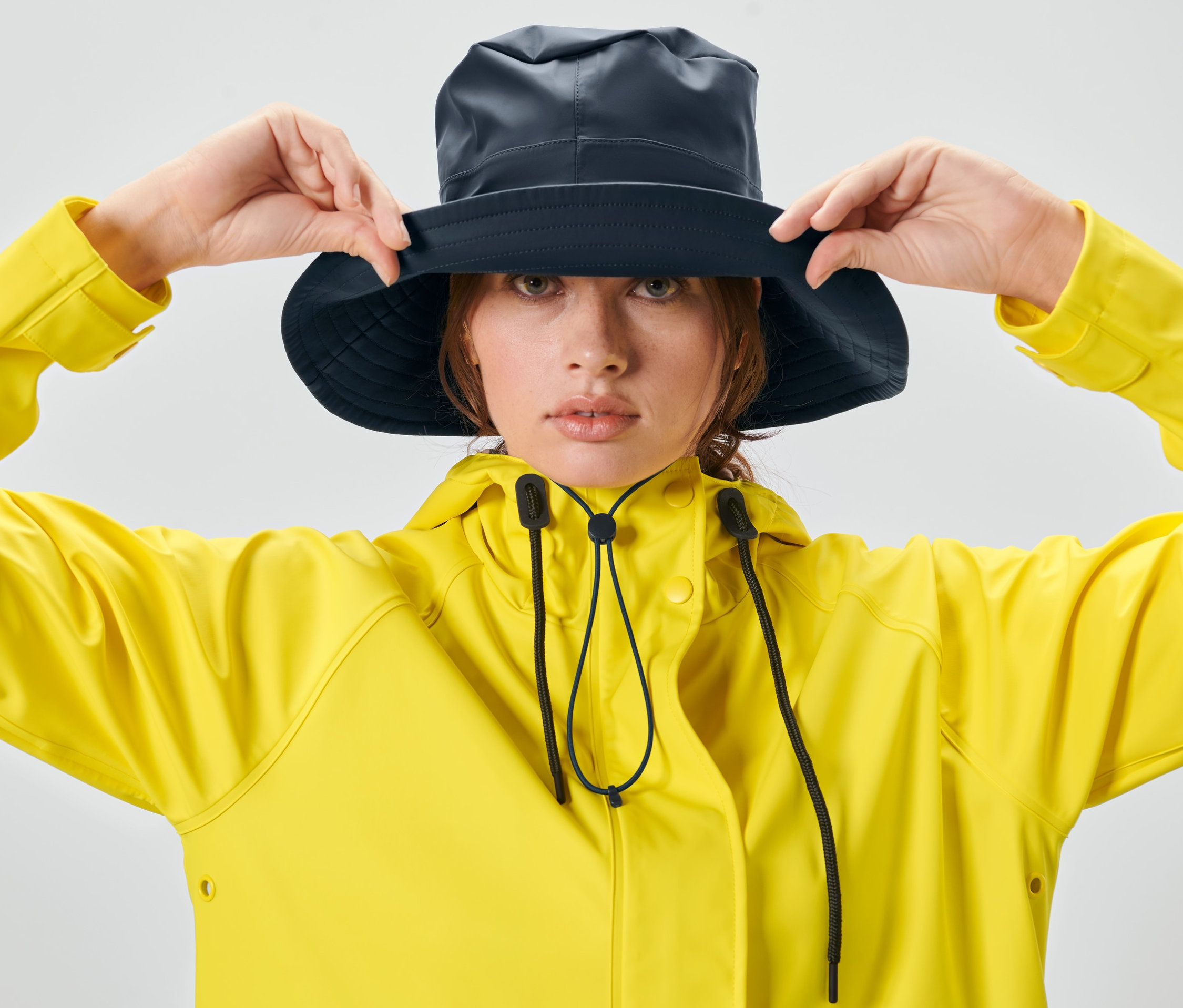 Žena si upravuje unisexový klobúk do dažďa a má na sebe žltú bundu.