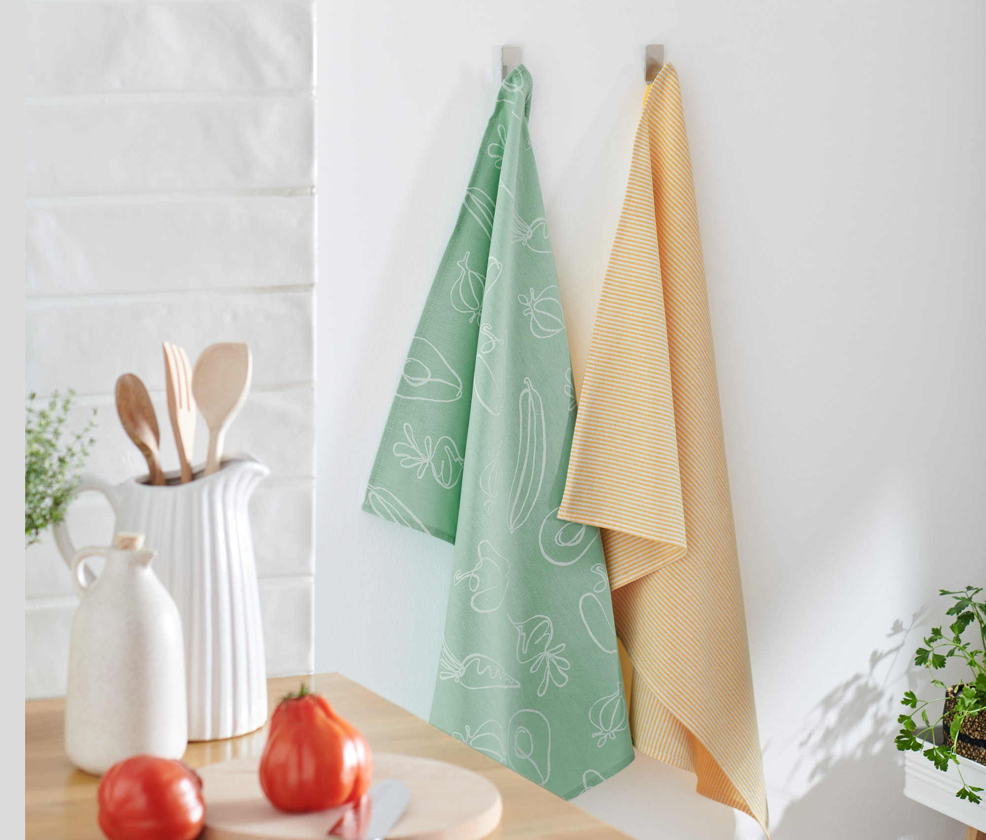Dve utierky na riad visia na stene, jedna svetlozelená s bielou potlačou zeleniny a druhá žltá s prúžkami. Utierky na riad, 2 ks