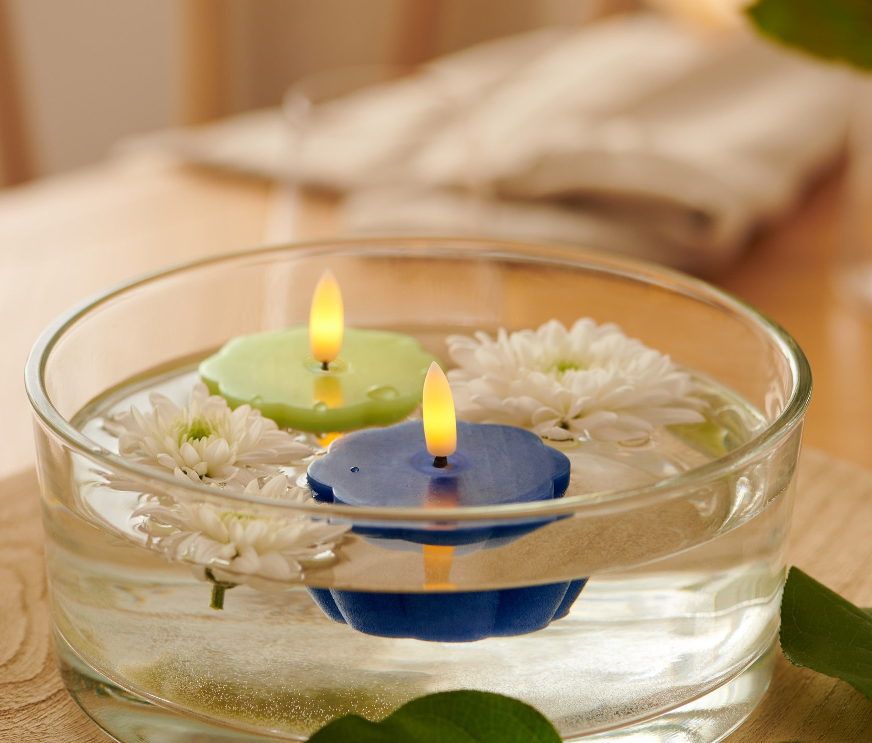 V priehľadnej miske plávajú dve LED čajové sviečky z pravého vosku, zelená a modrá, a biele kvety.