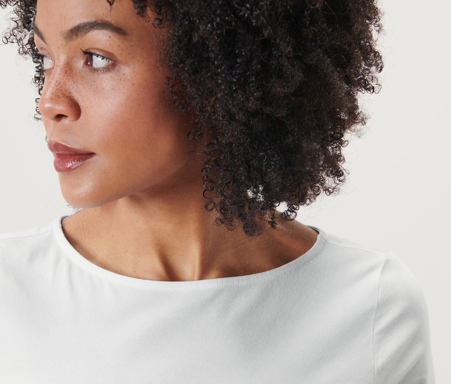Portrét ženy s hnedými kučeravými vlasmi, ktorá má na sebe biele tričko s trojštvrťovými rukávmi.