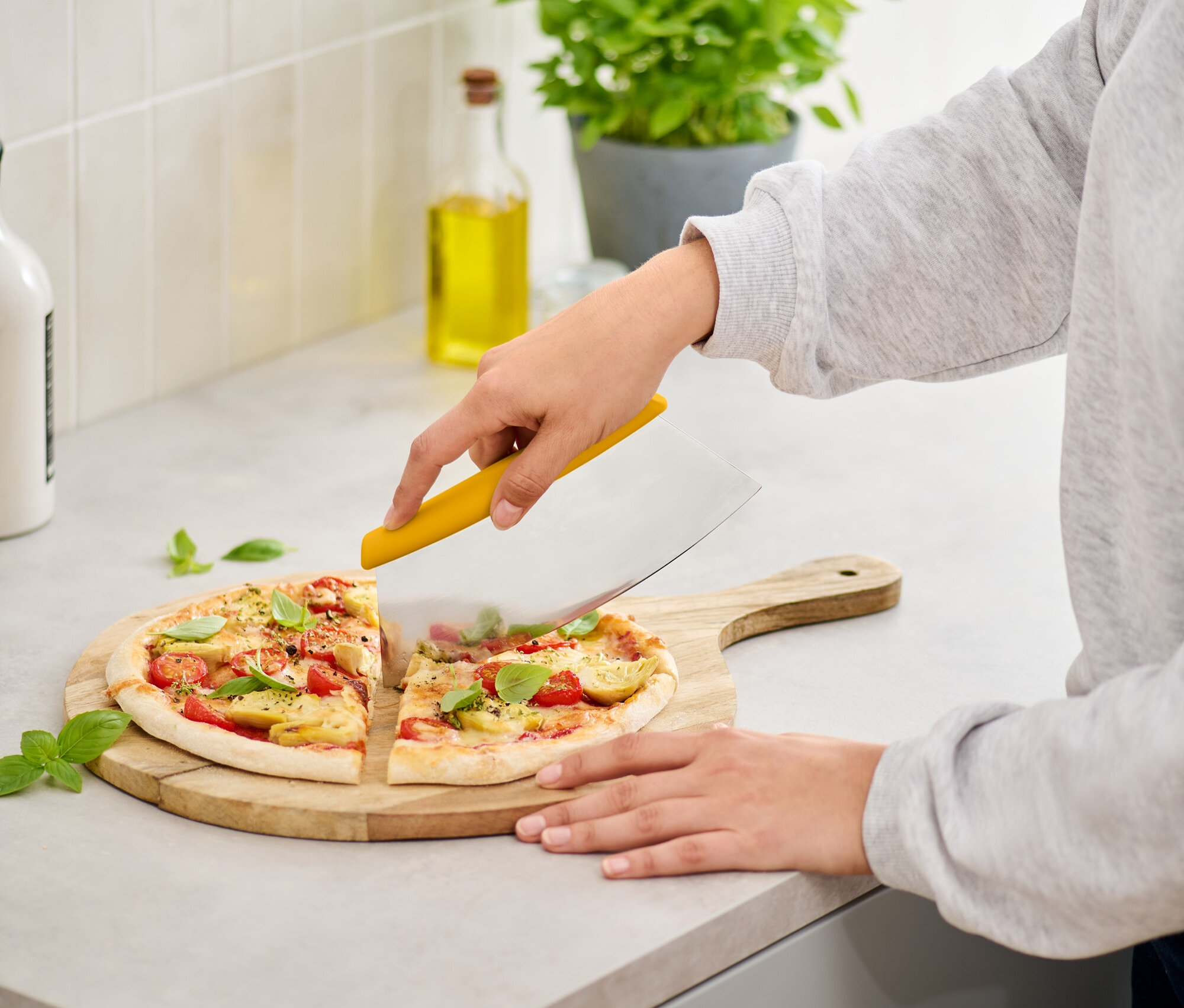 Žena krája pizzu s paradajkami a bazalkou na drevenej doske žltým krájačom na pizzu.