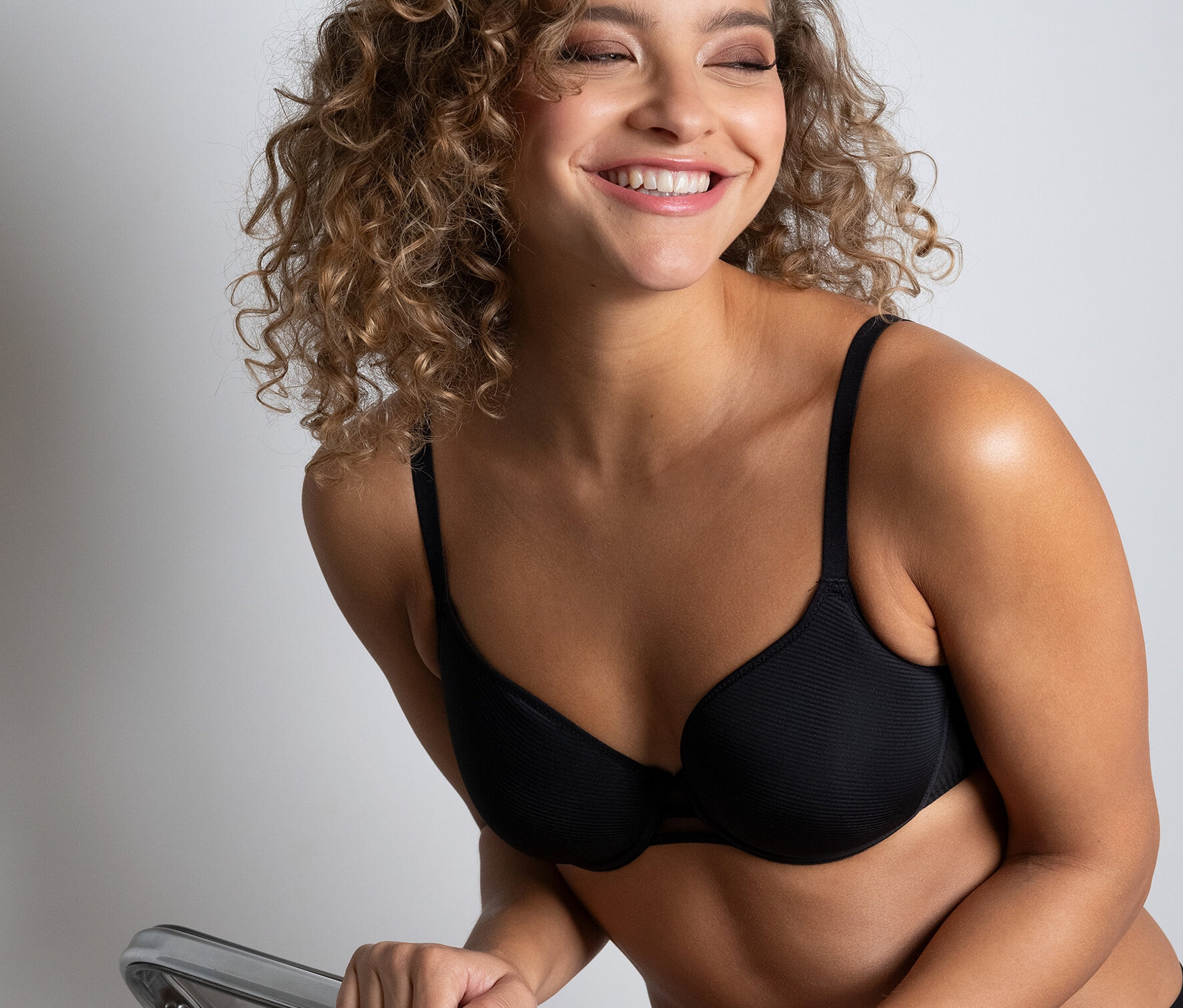Usmievajúca sa žena sedí na priehľadnej stoličke a má na sebe čiernu podprsenku Passionata »Nais«.