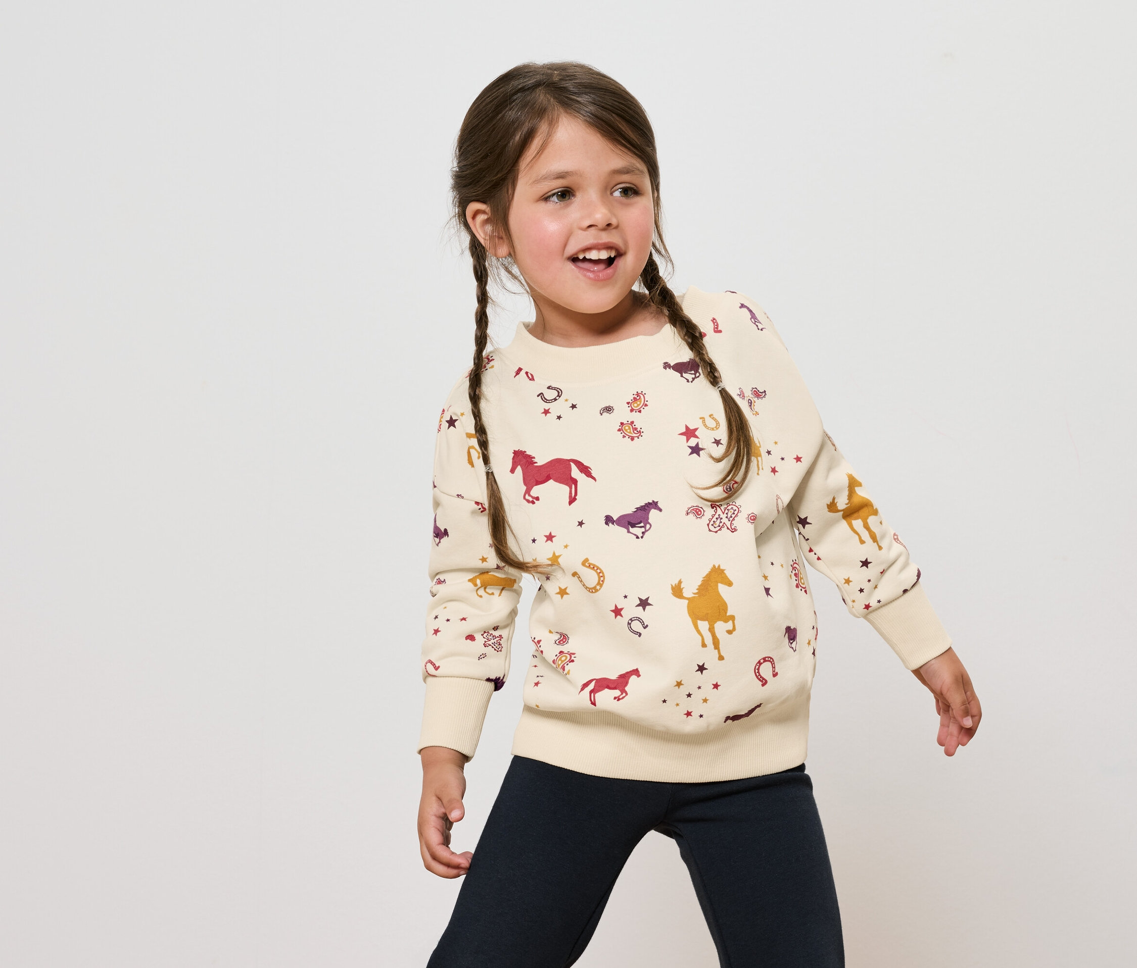 Dievča s hnedými vlasmi v pletených vrkočoch má na sebe krémovú mikinu s potlačou koní a tmavomodré nohavice.