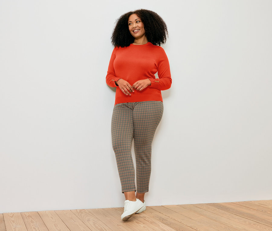 Žena pózuje v oranžovom svetri z jemnej pleteniny, vzorovaných nohaviciach a bielych topánkach.