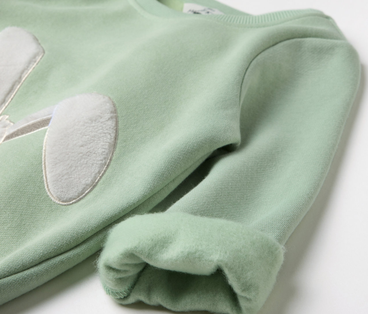 Svetlozelený pulóver s nášivkou králičích uší na hrudi a vyhrnutými rukávmi.