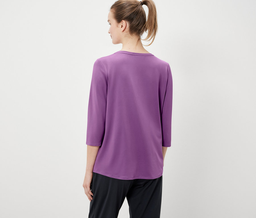 Zozadu je vidieť žena s vlasmi stiahnutými do copu, oblečená do fialového trička s trojštvrťovými rukávmi a čiernych nohavíc.