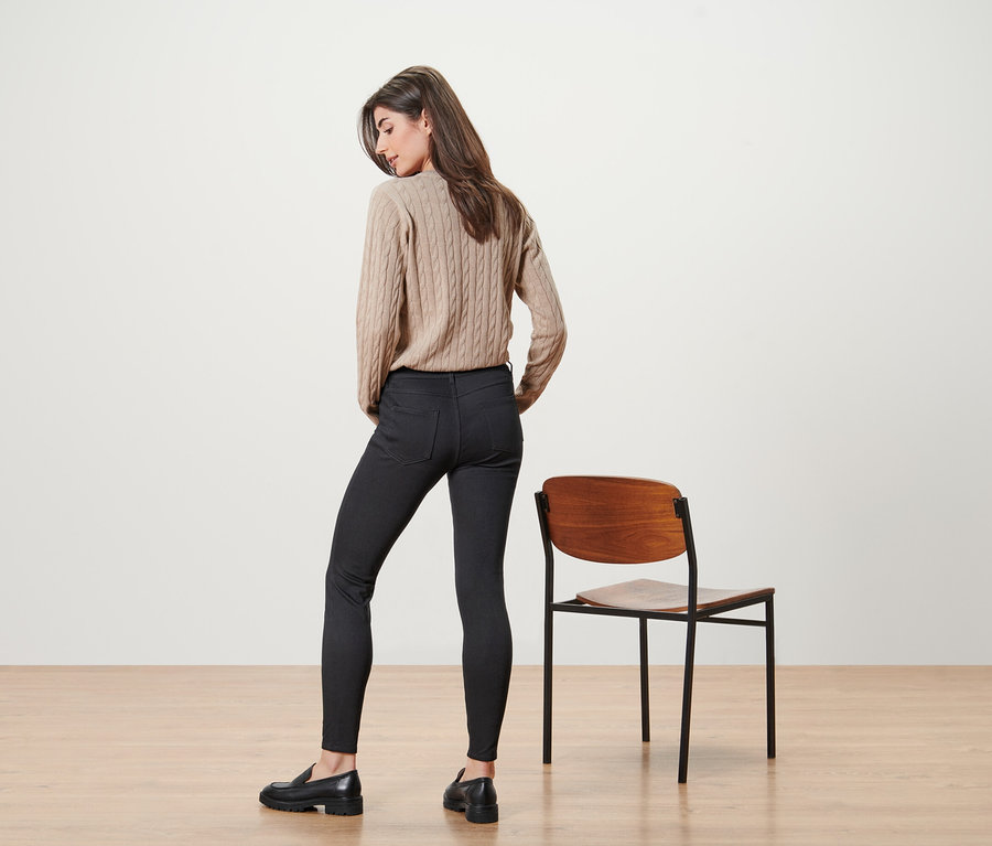 Žena pózuje v béžovom svetri a čiernych džínsoch vedľa stoličky.