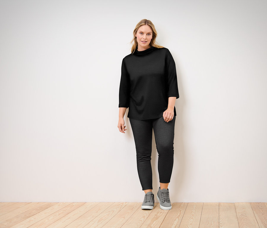 Žena pózuje v čiernom tope s trojštvrťovými rukávmi, čiernych džínsoch a sivých teniskách.