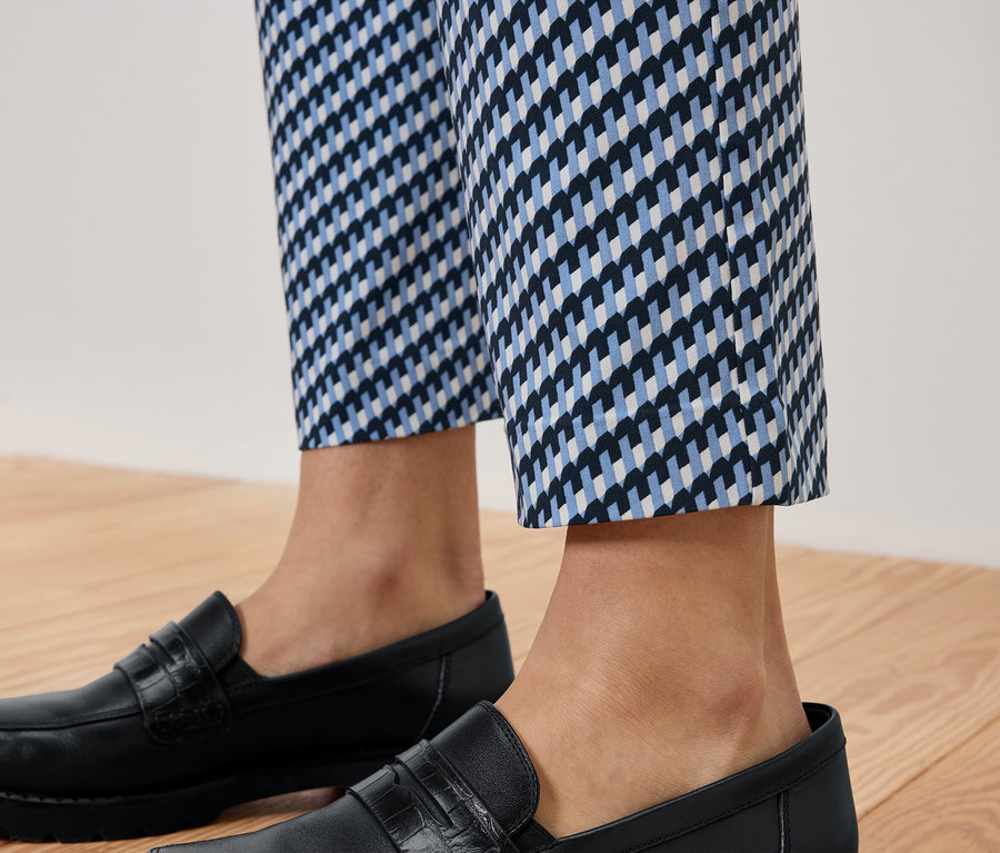 Nohy osoby v modro-bielych vzorovaných nohaviciach a čiernych kožených mokasínach na drevenej podlahe.