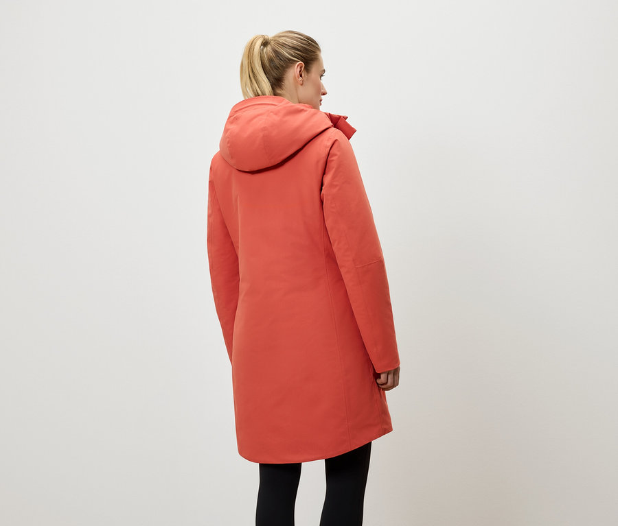 Žena chrbtom k fotoaparátu má na sebe koralovo červený kabát s kapucňou a čierne legíny.