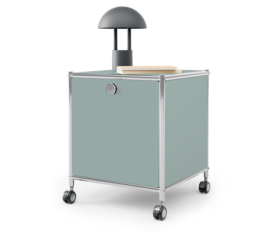 Sivá lampa a kniha na modrozelenom úložnom boxe na kolieskach.