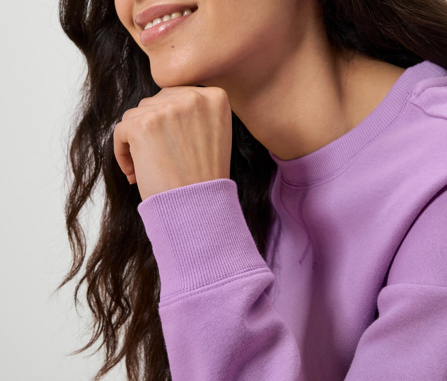 Detailný záber na ženu s vlnitými tmavými vlasmi, ktorá si rukou podopiera bradu. Má na sebe svetlofialovú mikinu.