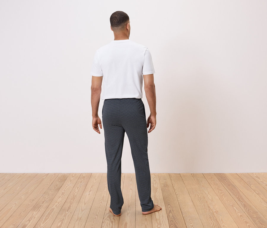 Muž chrbtom k fotoaparátu stojí na drevenej podlahe a má na sebe biele tričko a tmavé kockované nohavice.