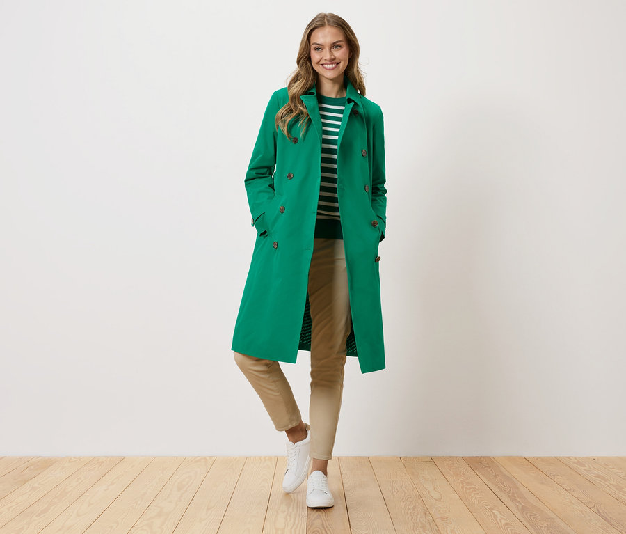 Žena v zelenom kabáte, pásikavom svetri, béžových nohaviciach a bielych teniskách stojí pred bielou stenou.