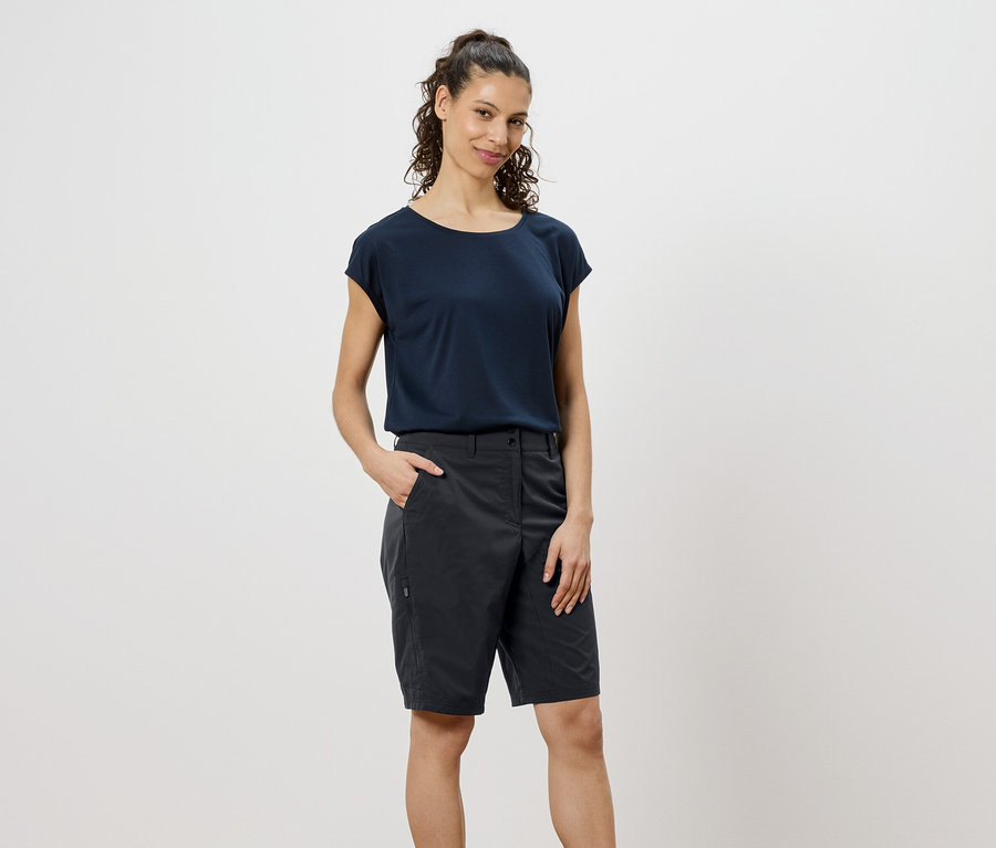 Žena s kučeravými hnedými vlasmi má na sebe tmavomodré tričko a sivé šortky, s rukou vo vrecku.