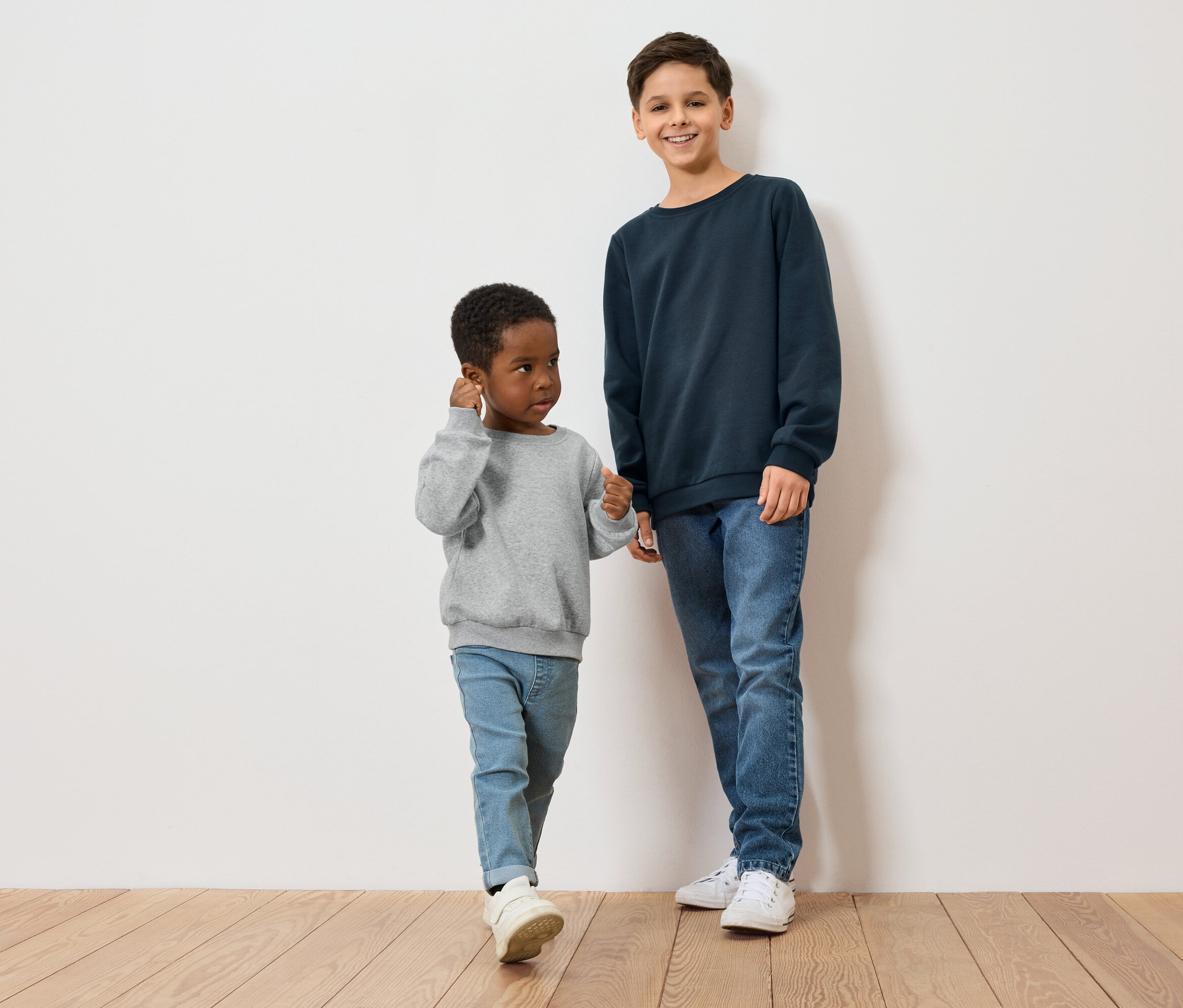 Dvaja chlapci, jeden s tmavou pokožkou v sivom svetri, druhý so svetlou pokožkou v modrom svetri, stoja na drevenej podlahe pred bielou stenou.