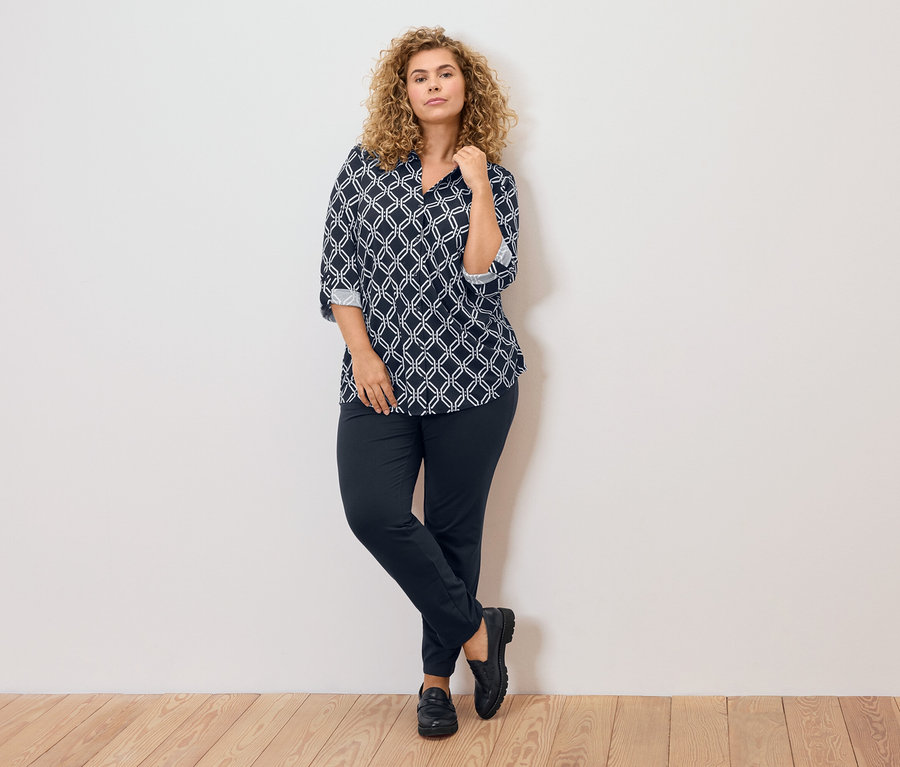 Žena s kučeravými blond vlasmi, oblečená v modro-bielej vzorovanej blúzke, tmavých nohaviciach a čiernych topánkach, opretá o stenu.