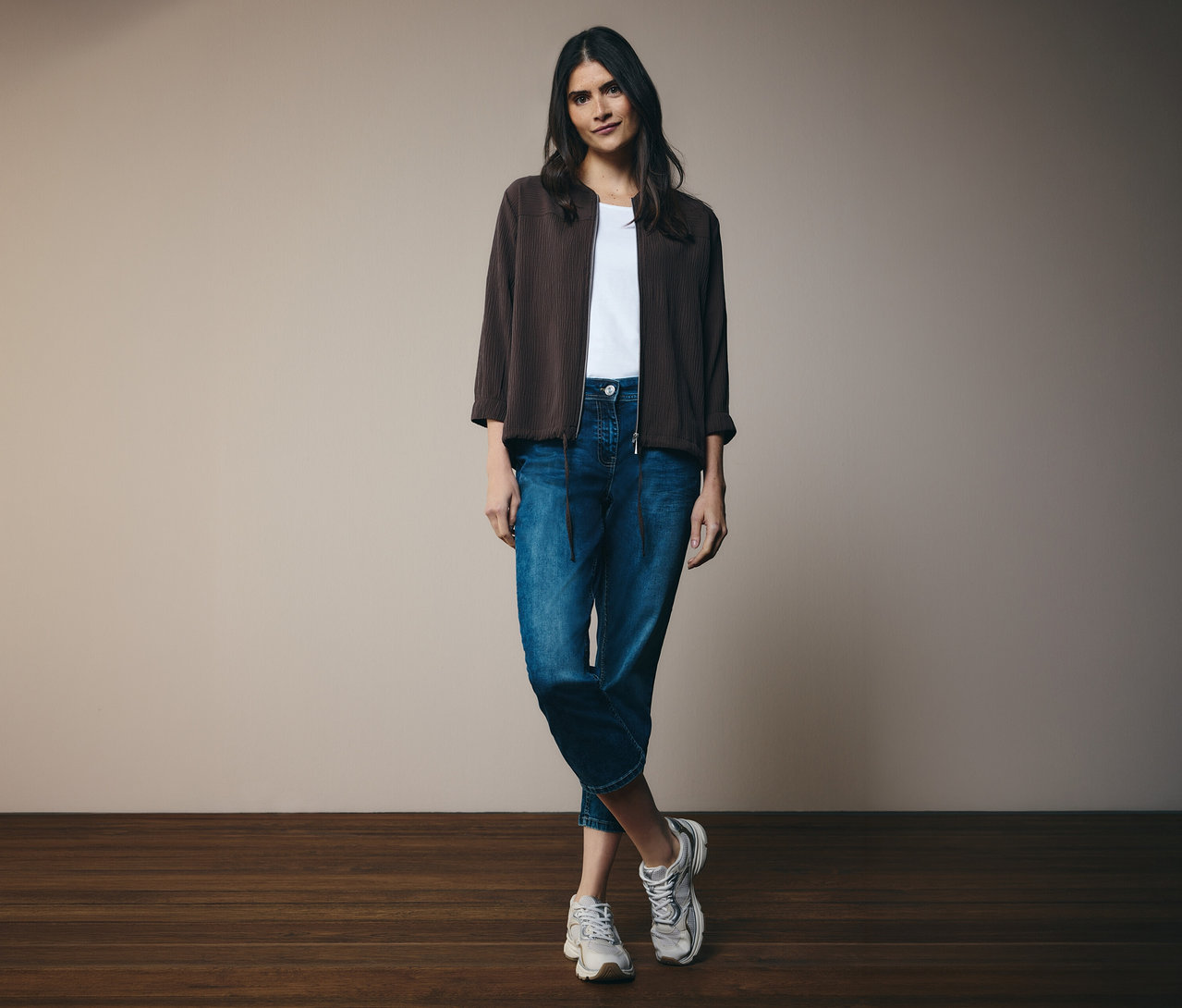 Žena v bielom tričku, džínsoch a hnedej bunde CECIL. Stojí pred stenou.