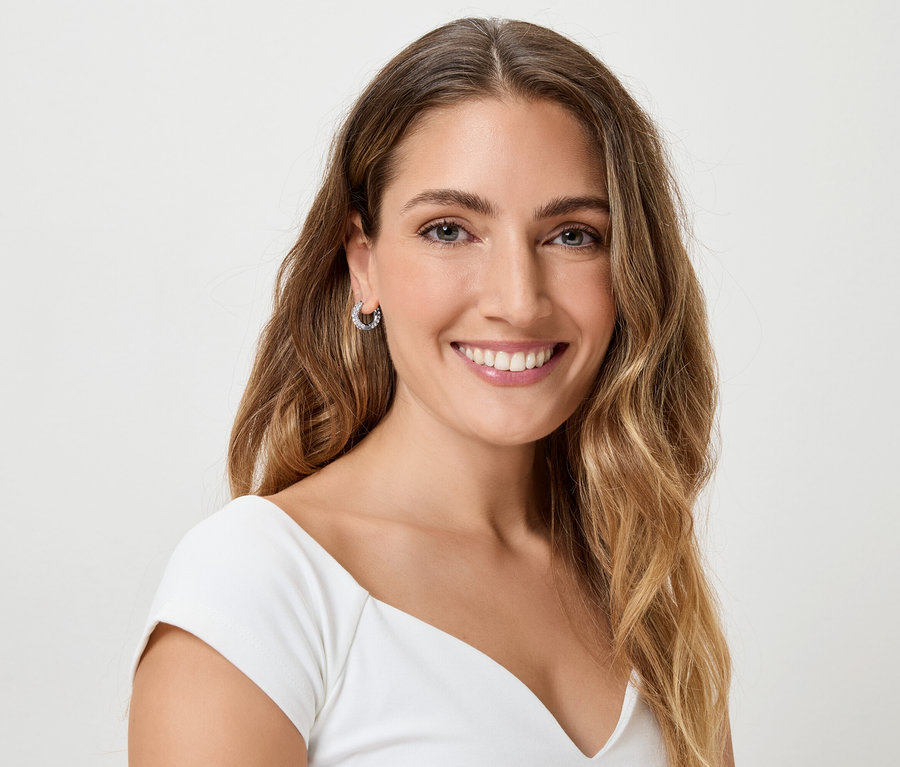 Žena s dlhými, vlnitými, hnedými vlasmi a náušnicami sa usmieva do kamery.