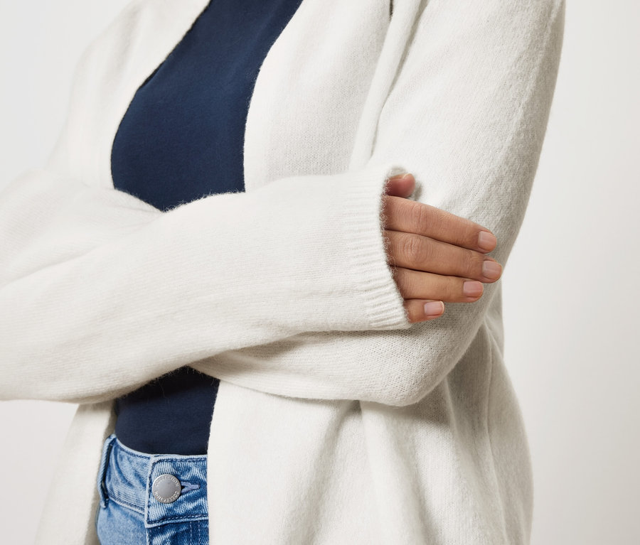 Žena so skríženými rukami má na sebe dlhý biely kardigán cez tmavomodrý top a modré džínsy.