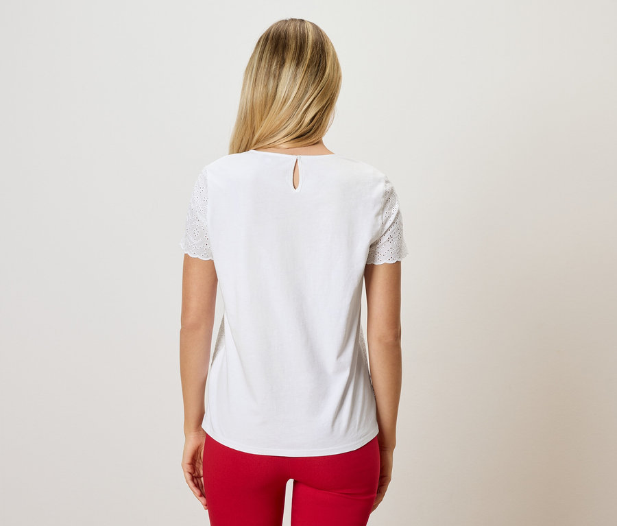 Pohľad zozadu na ženu s blond vlasmi v bielej blúzke s dierkovanou výšivkou na rukávoch a červených nohaviciach.