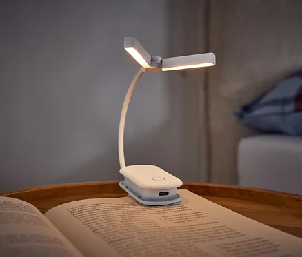 Biela klipová lampa na čítanie osvetľuje otvorenú knihu ležiacu na drevenom podnose. V pozadí je posteľ s károvaným vankúšom.