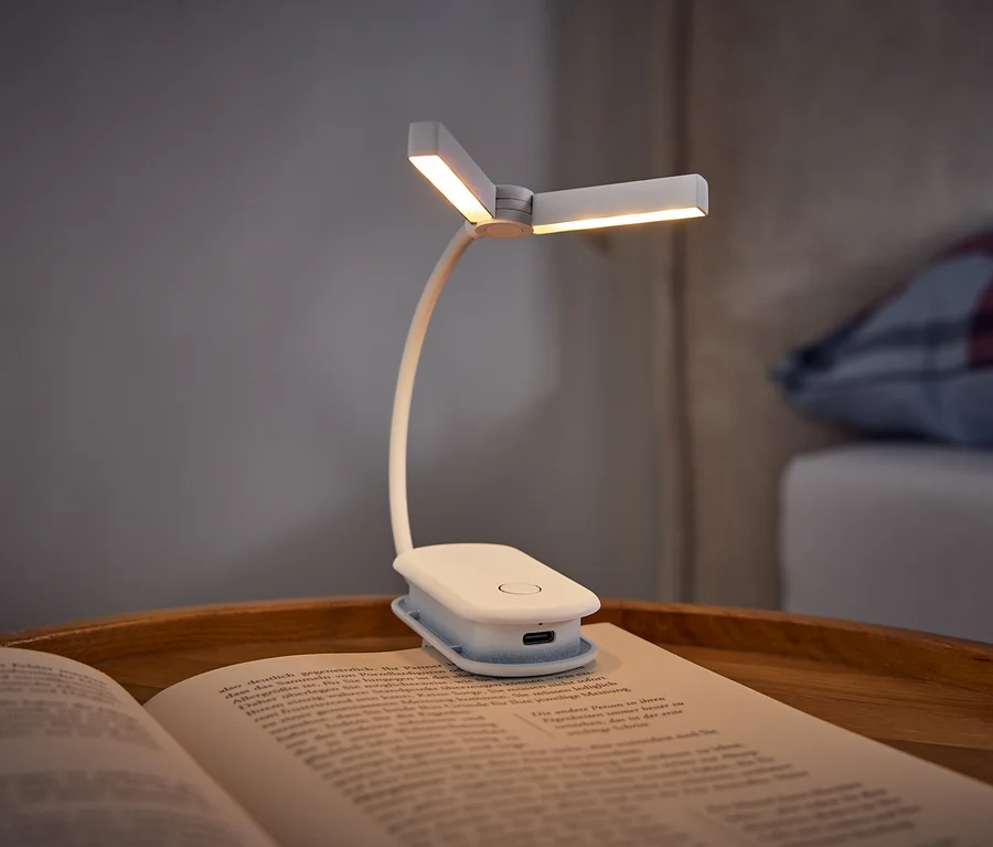 Biela klipová lampa na čítanie osvetľuje otvorenú knihu ležiacu na drevenom podnose. V pozadí je posteľ s károvaným vankúšom.