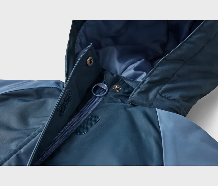 Detail modrej bundy zobrazujúci čiastočne rozopnutý zips, kapucňu, patentné gombíky a suché zipsy na chlopni proti vetru.
