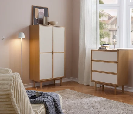 Svetlá obývačka s drevenou skrinkou Cabinetschrank s 2 dverami a komodou s 3 zásuvkami so svetlými čelami. Na komode stojí akumulátorová LED stolová lampa Eglo. Vľavo pohovka, vpravo okno so závesmi.
