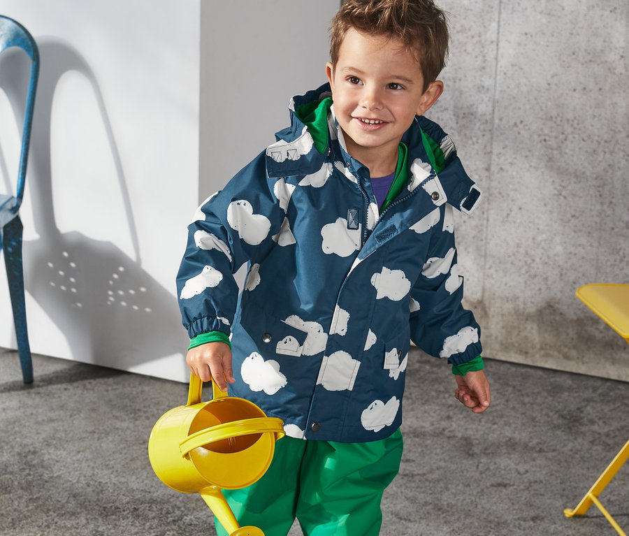 Chlapec drží žltú kanvicu na zalievanie a má na sebe bundu do dažďa z recyklovaného materiálu s obláčikmi, zelené nohavice a modré gumáky.