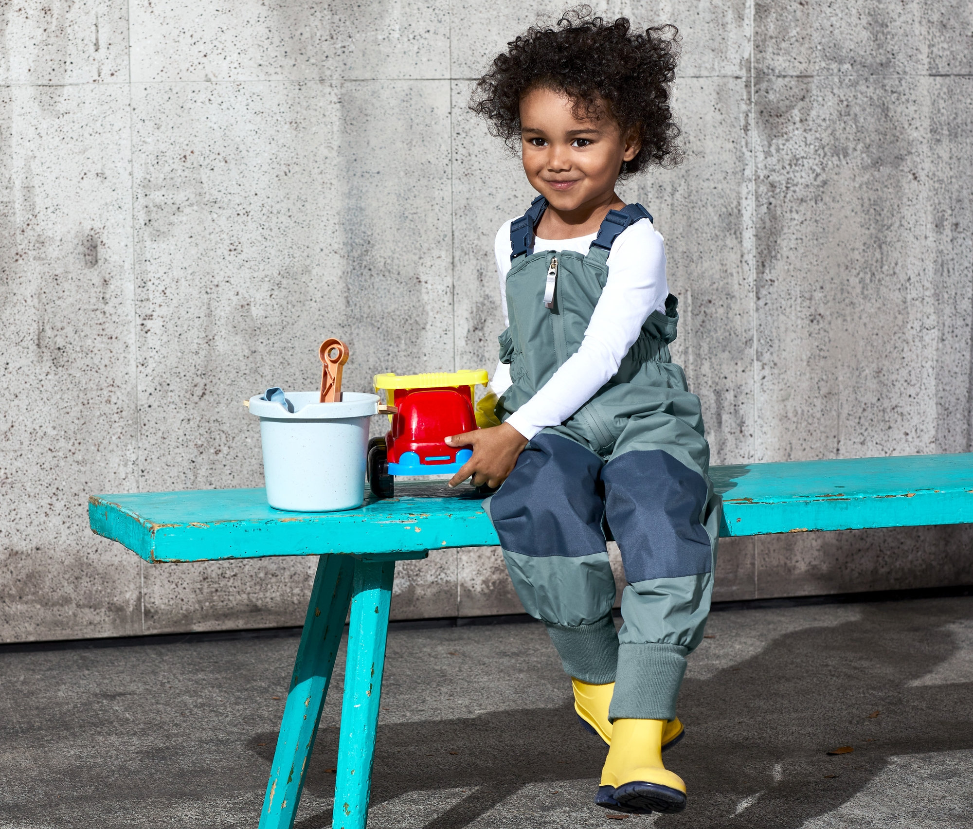 Dieťa sedí na lavičke a hrá sa s hračkami. Má na sebe nohavice do dažďa z recyklovaného materiálu, nastaviteľné.