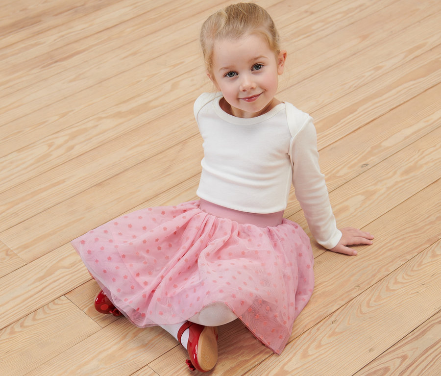 Usmievajúce sa dievčatko sedí na drevenej podlahe a má na sebe detskú tylovú sukňu.