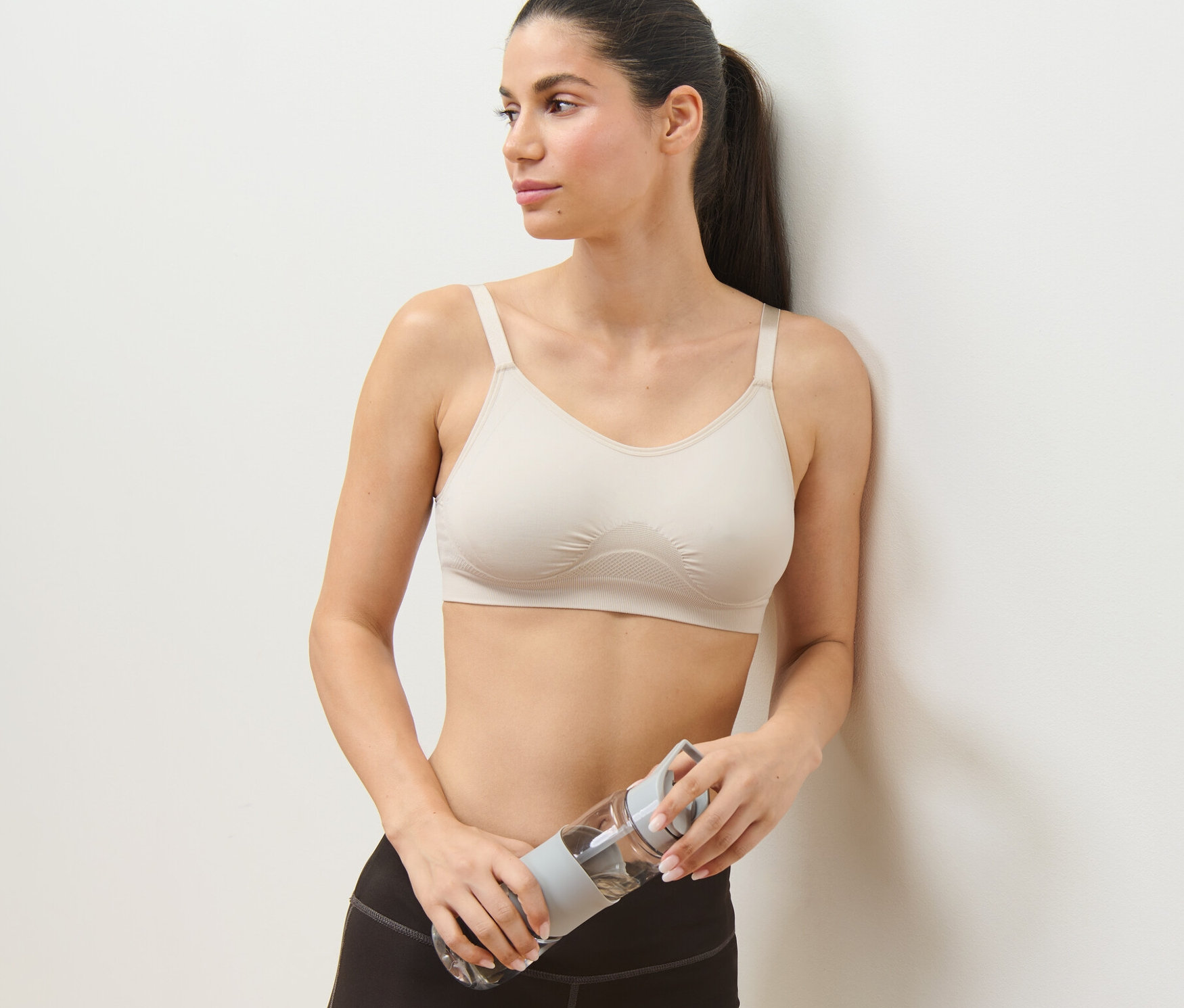 Žena opretá o stenu drží fľašu s vodou a má na sebe hnedú športovú podprsenku z 2-dielnej súpravy.