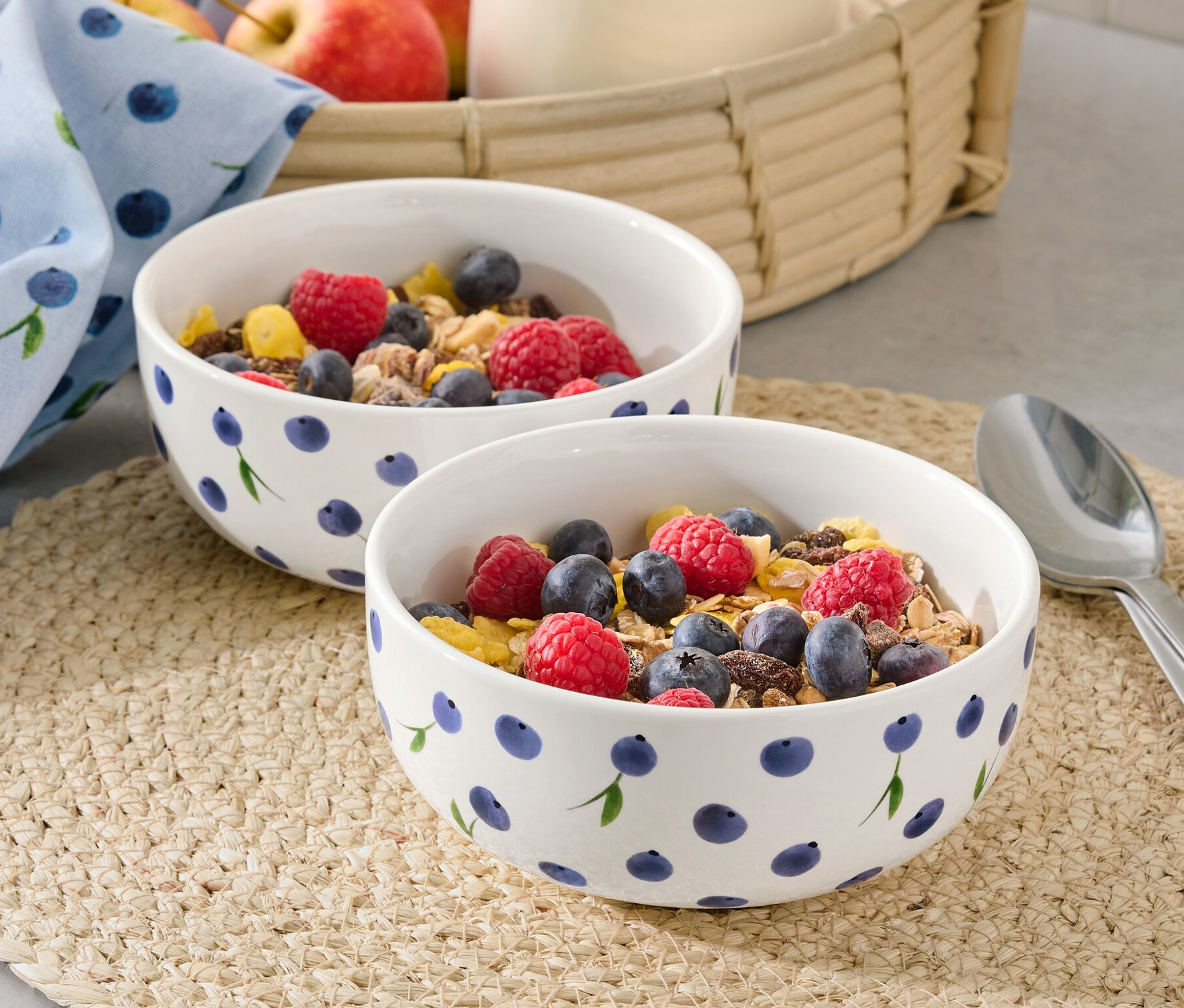 Dve misky s müsli, malinami a čučoriedkami na prestieraní. V pozadí džbán s mliekom a jablká v košíku.