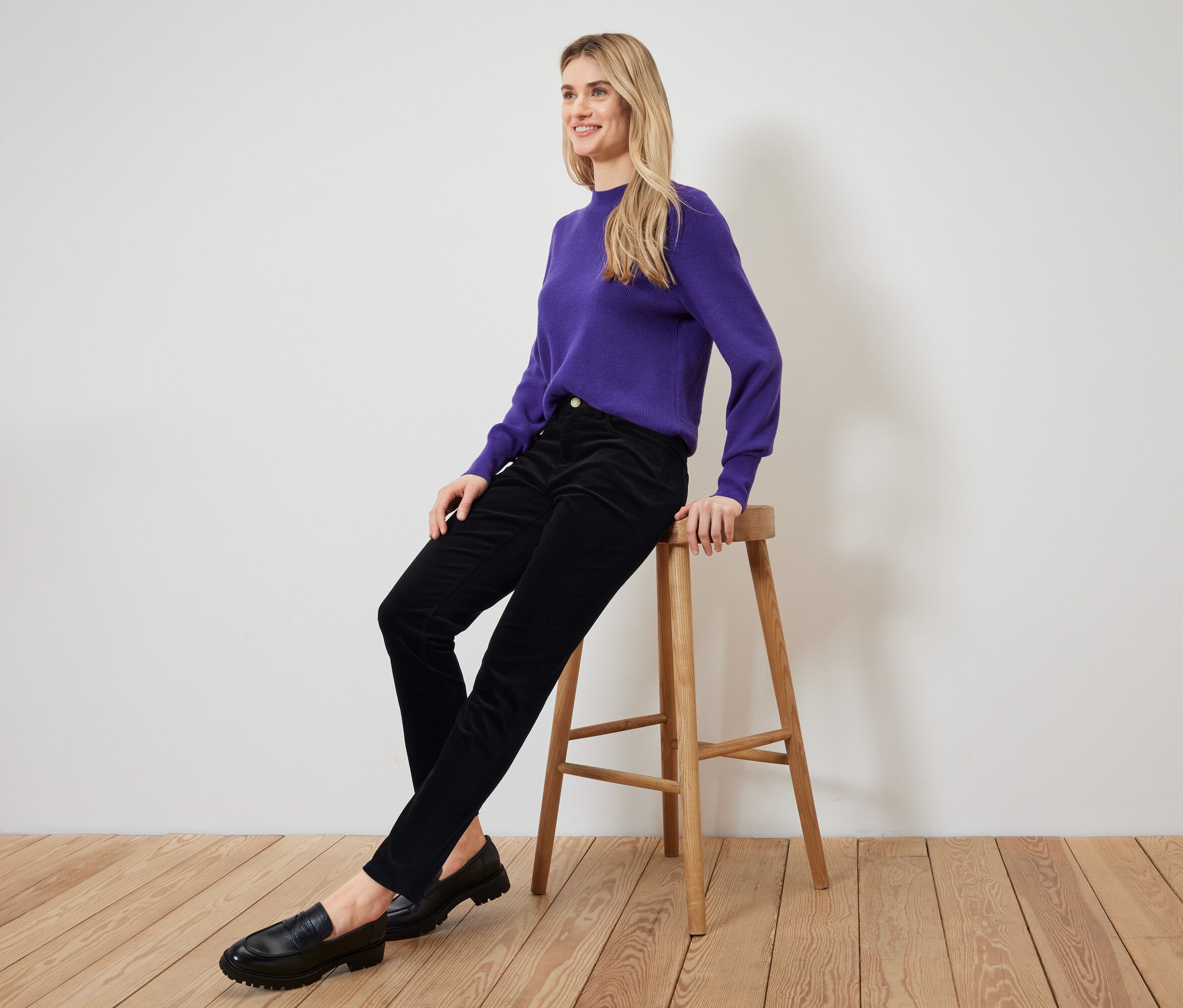 Blondínka sedí na drevenej stoličke, má na sebe fialový sveter, čierne nohavice a čierne mokasíny.