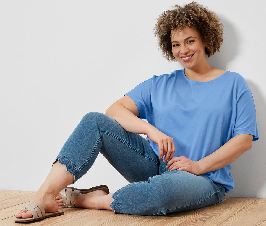 Žena sedí na drevenej podlahe a má na sebe svetlomodré blúzkové tričko a sedemosminové tregínsy s ažúrovou výšivkou.