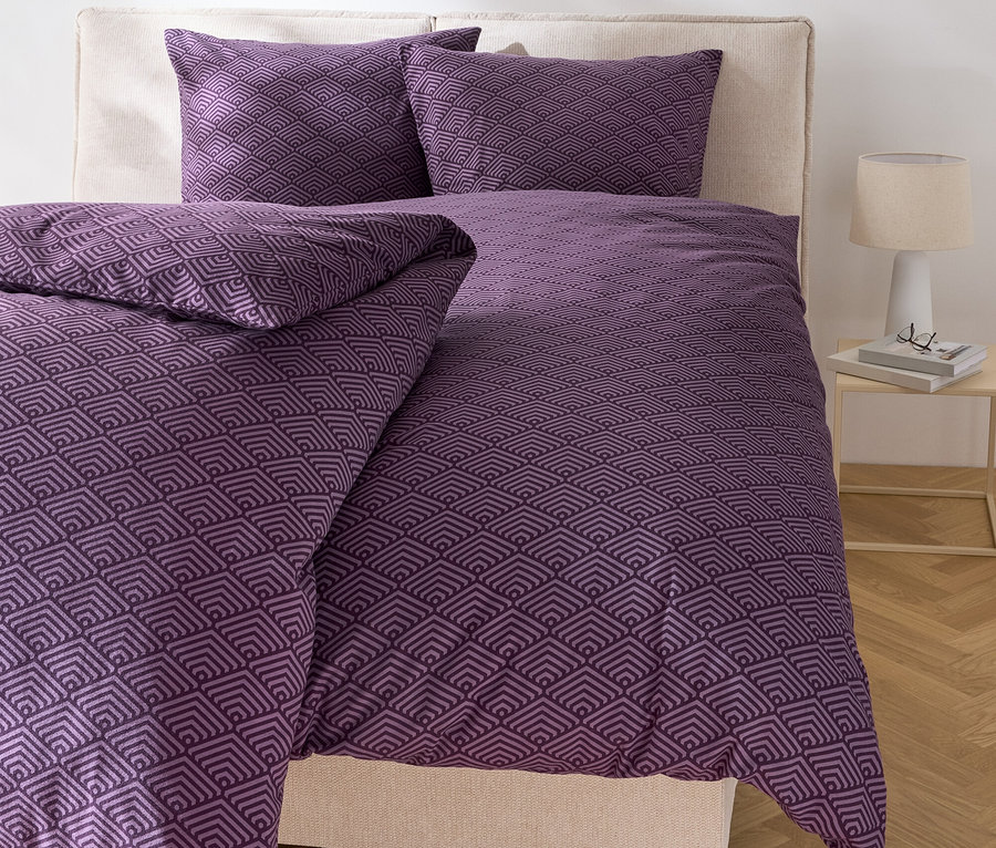 Posteľ s fialovou geometrickou posteľnou bielizňou a dvoma vankúšmi.
